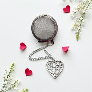 May include: A stainless steel tea infuser with a chain and a heart-shaped charm featuring floral cutouts. The infuser is round with a mesh basket. The image includes red heart-shaped paper cutouts and white flowers on a white background.