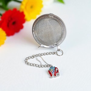 May include: A stainless steel tea infuser with a chain and a decorative charm. The charm is an elephant design with red enamel and colorful rhinestones. The infuser is round and has a fine mesh.