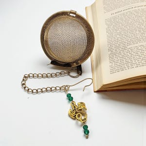 May include: A vintage-style tea infuser with a mesh basket and a decorative chain. The infuser features a gold-colored dragon charm with green beads. The infuser is made of metal and is designed for steeping loose leaf tea.