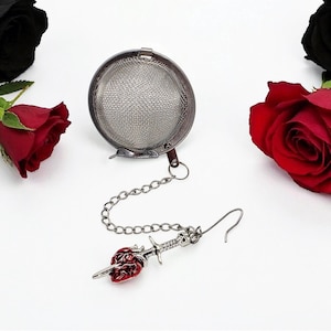 May include: A stainless steel tea infuser with a chain and a decorative charm. The charm is a red rose pierced by a sword with a skull on the hilt. The infuser is round and has a fine mesh. Red and black roses are in the background.
