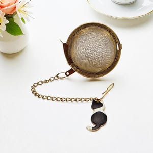 May include: A round, antique brass tea infuser with a chain and a black and white cat charm. The infuser is open, revealing a fine mesh interior. The charm is a silhouette of a cat. The tea infuser is designed for steeping loose leaf tea.