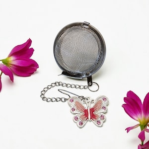 Op de afbeelding: Een roestvrijstalen thee-ei met een vlinderbedel. De infuser is een ronde mesh bal met een scharnierende sluiting. De vlinderbedel is zilverkleurig met roze emaille en roze kristallen accenten. De thee-infuser is bevestigd aan een ketting.