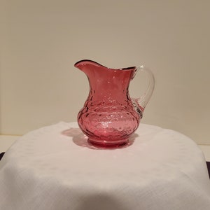 May include: A small, pink glass pitcher with a clear glass handle. The pitcher has a textured, bubble-like design.