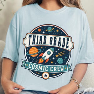 May include: A light blue t-shirt featuring a circular graphic with a space theme. The design includes a rocket, planets, stars, and the text "THIRD GRADE" and "COSMIC CREW" in a bold, dark blue font. The shirt is labeled as chambray color.
