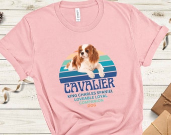 Puede incluir: Camiseta rosa con un gráfico de puesta de sol retro y un perro Cavalier King Charles Spaniel. El texto "Cavalier" está en azul con las palabras "King Charles Spaniel, Adorable, Leal, Compañero Perro" en negro debajo.