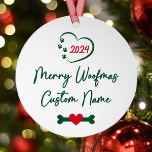 Pu&ograve; includere: Una decorazione natalizia rotonda bianca con un nastro rosso. L'ornamento presenta un cuore verde con impronte di zampe e l'anno 2024. Il testo recita "Merry Woofmas Custom Name". Un design a forma di osso e cuore &egrave; in basso.