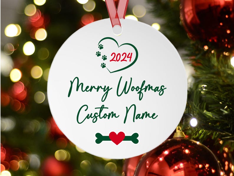 Pu&ograve; includere: Una decorazione natalizia rotonda bianca con un nastro rosso. L'ornamento presenta un cuore verde con impronte di zampe e l'anno 2024. Il testo recita "Merry Woofmas Custom Name". Un design a forma di osso e cuore &egrave; in basso.