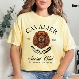 May include: Butter-colored t-shirt featuring a Cavalier King Charles Spaniel graphic. The design includes the text "CAVALIER Social Club" with "EST 2023" and "BRIGHTON MICHIGAN" in a classic font. The shirt is a relaxed fit.