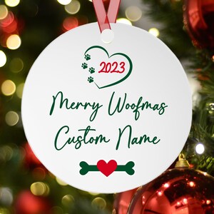 May include: White round Christmas ornament with a green heart containing the text "2023" in red. The text "Merry Woofmas Custom Name" is written in green cursive script. A green bone with a red heart in the center is at the bottom of the ornament.