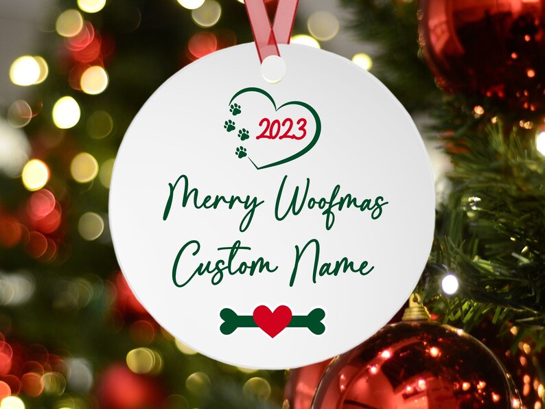 May include: White round Christmas ornament with a green heart containing the text "2023" in red. The text "Merry Woofmas Custom Name" is written in green cursive script. A green bone with a red heart in the center is at the bottom of the ornament.