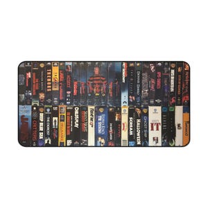 Puede incluir: Una alfombrilla de escritorio rectangular con una colección de cintas VHS de películas de terror. Los títulos incluyen "Halloween", "Scream", "The Shining" y "Friday the 13th". La alfombrilla es negra con esquinas redondeadas.