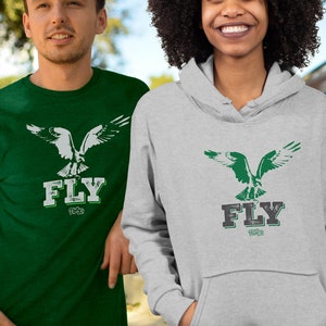 Könnte beinhalten: Zwei Personen tragen Shirts mit einer grünen Seeadler-Grafik und dem Wort "FLY" in weißen Buchstaben. Das Shirt links ist grün und das Shirt rechts ist grau.