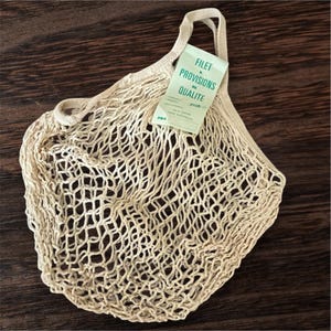 May include: A beige, open-weave shopping bag with a handle and a tag that reads "FILET & PROVISIONS DE QUALITE" in green text. The bag is made of a woven material and is sitting on a dark brown wooden surface.