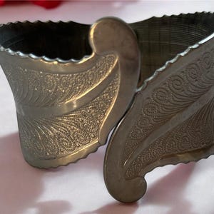 May include: A silver metal bracelet with an intricate floral design. The bracelet is adjustable and has a wide, open design.