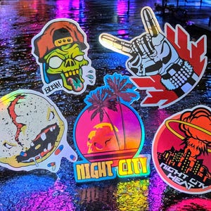 May include: A selection of holographic stickers with bold designs. The stickers depict a zombie wearing a cap, a skeleton hand in a rock on gesture, a skull with a sunset and palm trees, and a cityscape with an atomic explosion. The text "Night City" and "Night City Heat" are visible.