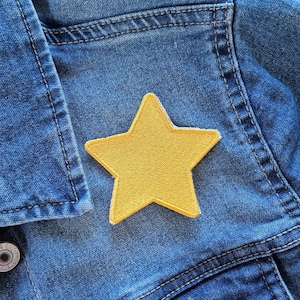 Könnte beinhalten: Nahaufnahme einer blauen Jeansjacke mit einem leuchtend gelben sternförmigen Aufnäher. Die Jacke hat sichtbare Nähte und Metallknöpfe. Der Stern-Aufnäher befindet sich mittig auf der Vorderseite der Jacke.