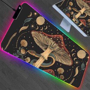 May include: Black rectangular gaming mouse mat with a colourful LED edge. The mat features a detailed illustration of a mushroom, snake, moons, and stars. The design is in shades of brown, red, and gold.