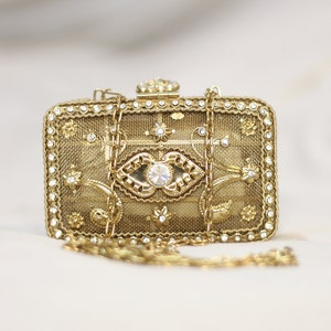 Puede incluir: Un bolso de mano rectangular dorado con una cadena. El bolso presenta intrincados diseños florales y geométricos, y está adornado con pequeños y brillantes pedrería. El cierre está adornado con una gran pedrería transparente.
