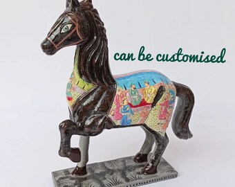 Puede incluir: Una figurita de caballo de madera pintada a mano con detalles intrincados. El caballo es marrón con una silla de montar y jinetes coloridos. La base es gris con un patrón decorativo.