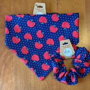 May include: A blue bandana and scrunchie set featuring a red apple and white polka dot design. The handmade accessories have a tag that reads "Handmade with love". The bandana is triangular, and the scrunchie is circular.