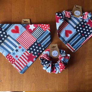 May include: Three patriotic-themed pet accessories: two bandanas and a scrunchie. The bandanas feature a patchwork design with red hearts, white stars, and blue and white stripes. The scrunchie has a matching pattern. All items have a tag.