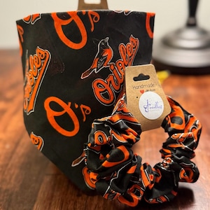 Op de afbeelding: Zwarte en oranje Baltimore Orioles bandana en scrunchie set. De bandana en scrunchie zijn voorzien van het Orioles logo en vogelafbeelding. De scrunchie staat naast een handgemaakt label.
