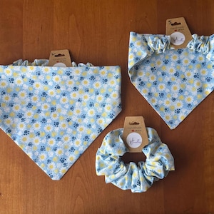 Puede incluir: Juego de tres accesorios para mascotas con tela azul claro con estampado de margaritas y huellas de patas. Incluye dos bandanas y una scrunchie. Cada artículo tiene una etiqueta marrón con un logotipo manuscrito.