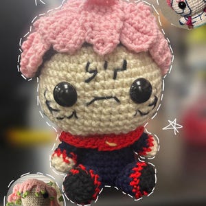 May include: A handmade crochet doll with a light beige face, large black eyes, and a pink, petal-like head covering. The doll wears a navy blue outfit with red accents and black feet. Additional smaller crochet dolls are also shown.