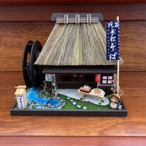 Könnte beinhalten: Ein japanisches Miniaturhaus mit Strohdach, einem Wasserrad und einem kleinen Garten. Das Haus hat ein Schild mit der Aufschrift "松茸ごはん" in japanischer Sprache.