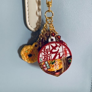 Könnte beinhalten: Ein goldfarbener Schlüsselanhänger mit zwei Anhängern. Ein Anhänger ist ein Miniatur-Gebäck, rund, gelb und orange. Der andere ist ein roter und weißer Miniatur-Lebensmittelbehälter mit japanischem Text und einem Bild von Nudeln.
