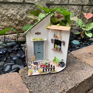 Könnte beinhalten: Ein Miniatur-Weißes Haus mit einem grünen Dach, einer blauen Tür und einem kleinen Fenster. Das Haus ist mit Miniaturpflanzen, Blumen und einem kleinen Stuhl dekoriert. Das Haus hat die Nummer 2120 über der Tür.