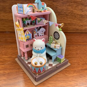 May include: A miniature dollhouse with a pink and white color scheme. The dollhouse features a bookshelf with a variety of miniature items, including a toy rocking horse, a book, and a toy dog. A white polar bear doll is playing a drum set in front of the bookshelf.