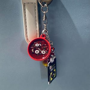 Könnte beinhalten: Ein roter Schlüsselanhänger in Form einer Suppenschüssel mit weißen und grünen Details. Der Anhänger ist an einer silbernen Kette befestigt, an der ein kleines Etikett mit japanischem Text und einem gelb-schwarzen Design hängt.