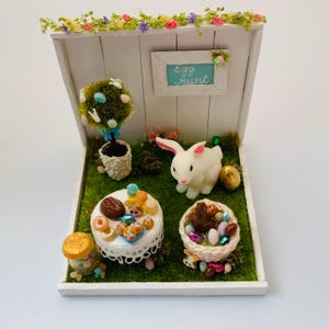 Puede incluir: Una escena de Pascua en miniatura con un conejito blanco, una mesa con golosinas, una cesta de huevos y un cartel que dice "Búsqueda de huevos".