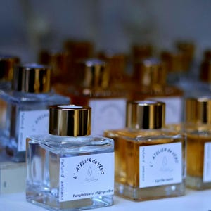 Peut inclure: Une collection de flacons de parfum carrés en verre avec des bouchons dorés, chacun portant une étiquette blanche avec le texte L'ATELIER de VERO, une icône de goutte et des noms de senteurs comme Pamplemousse et gingembre ou Vanille noire.