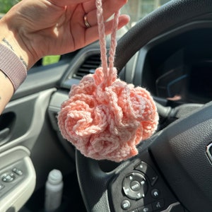 May include: A light pink crocheted bath pouf featuring a dense, ruffled texture and a matching crocheted loop handle for hanging. The item is constructed from soft yarn with a circular, layered design that creates a voluminous, flower-like appearance.