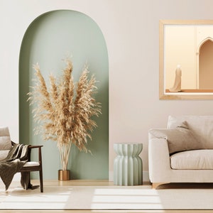 Puede incluir: Un interior de sala de estar con un sof&aacute; beige claro, un sill&oacute;n de madera con una manta de lana marr&oacute;n, una mesa auxiliar verde claro y una impresi&oacute;n enmarcada de una persona con una t&uacute;nica blanca caminando a trav&eacute;s de un arco.