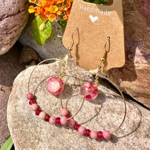 Op de afbeelding: Gouden creolen met roze kralen en een roze bloem ingekapseld in hars die aan elke creool hangt. De oorbellen zijn handgemaakt en bevinden zich op een bruine kaart met het woord "Handmade" erop.