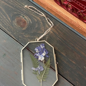 Op de afbeelding: Een goudkleurige, geometrisch gevormde glazen ornament met een geperste blauwe en witte bloem en groene varen. Het ornament hangt aan een natuurlijke touwtje. De achtergrond is een donker houten oppervlak.