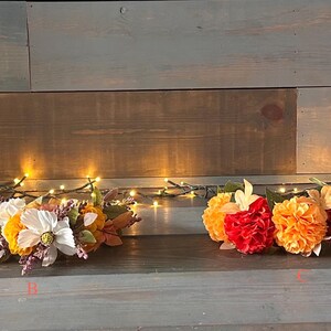 Può includere: Quattro fasce floreali nei toni dell'arancione, del rosso, del giallo e del bianco sono esposte su una superficie di legno. Le fasce sono adornate con vari tipi di fiori artificiali. Una fila di piccole luci dai toni caldi è dietro le fasce.