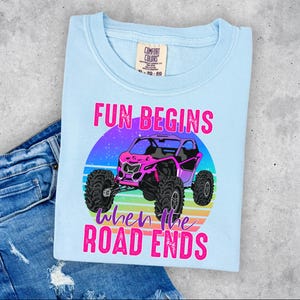 May include: Light blue t-shirt with a graphic of a pink off-road vehicle. The design includes the text "FUN BEGINS when the ROAD ENDS" in pink. The shirt is folded on top of a pair of blue denim jeans.