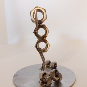 Puede incluir: Una escultura de metal plateado de una persona levantando pesas. La escultura est&aacute; hecha de tuercas y tornillos y est&aacute; de pie sobre una base redonda.