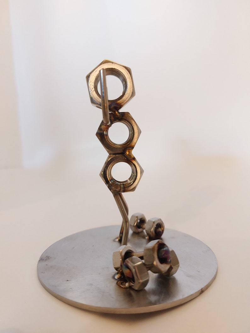 Puede incluir: Una escultura de metal plateado de una persona levantando pesas. La escultura est&aacute; hecha de tuercas y tornillos y est&aacute; de pie sobre una base redonda.