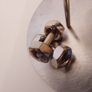 Puede incluir: Tres tuercas de metal plateado apiladas una encima de la otra, con una varilla de metal que pasa por el centro de las tuercas. Las tuercas est&aacute;n sobre una superficie de metal plateado.