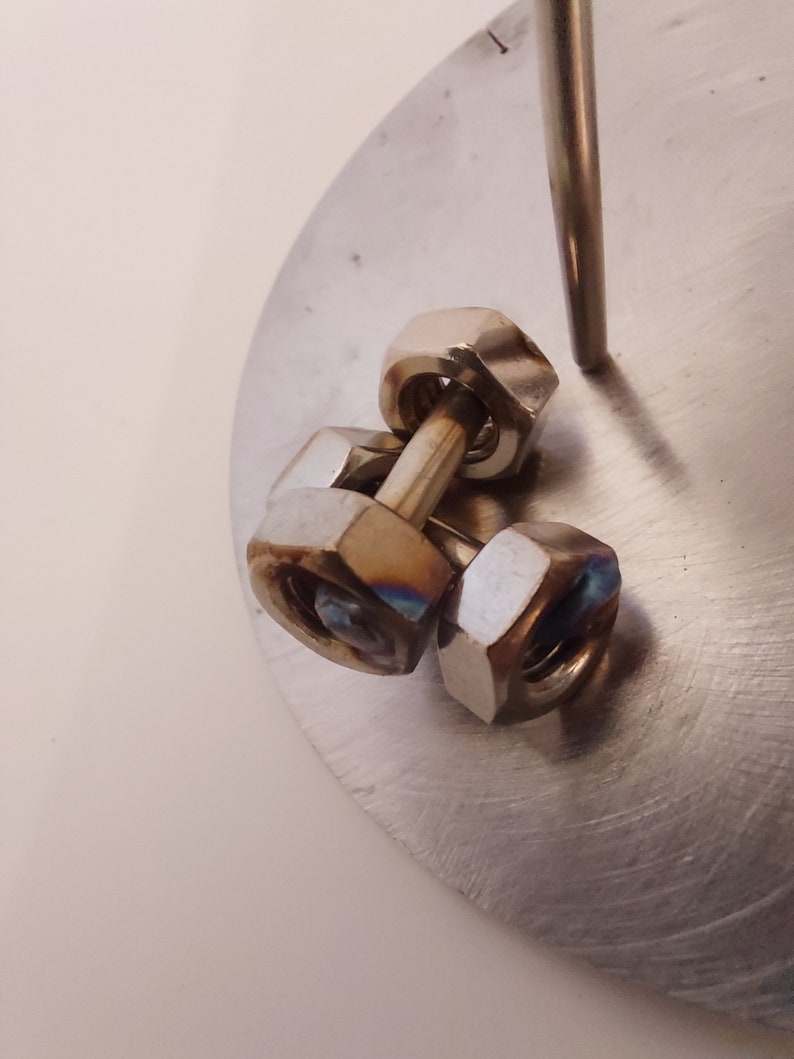 Puede incluir: Tres tuercas de metal plateado apiladas una encima de la otra, con una varilla de metal que pasa por el centro de las tuercas. Las tuercas est&aacute;n sobre una superficie de metal plateado.