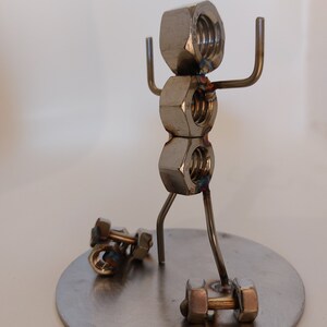 Puede incluir: Una escultura de metal de una persona levantando pesas. La persona est&aacute; hecha de tuercas y tornillos, y las pesas tambi&eacute;n est&aacute;n hechas de tuercas y tornillos. La escultura est&aacute; de pie sobre una base redonda de metal.