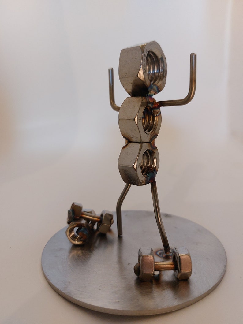Puede incluir: Una escultura de metal de una persona levantando pesas. La persona est&aacute; hecha de tuercas y tornillos, y las pesas tambi&eacute;n est&aacute;n hechas de tuercas y tornillos. La escultura est&aacute; de pie sobre una base redonda de metal.