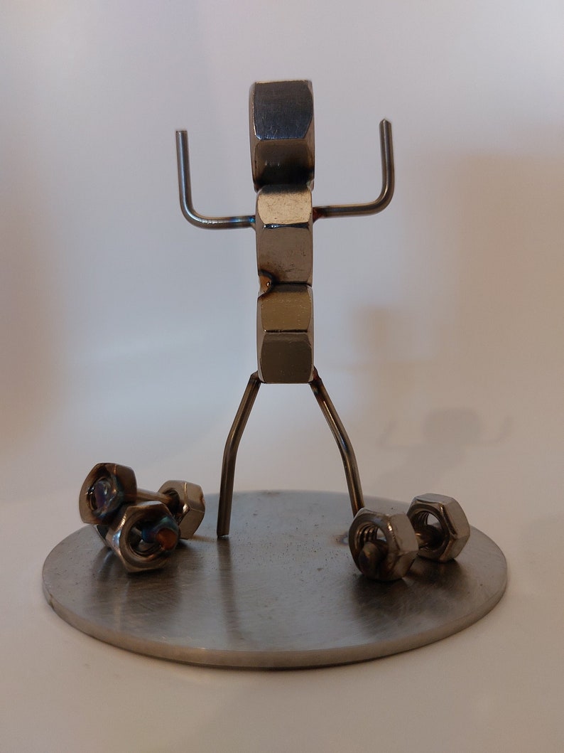 Puede incluir: Una escultura de metal de una persona levantando pesas. La persona est&aacute; hecha de tuercas y tornillos y est&aacute; de pie sobre una base redonda de metal. Las pesas tambi&eacute;n est&aacute;n hechas de tuercas y tornillos.