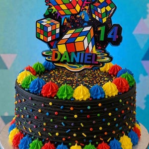 May include: A black frosted cake decorated with colorful frosting swirls and sprinkles. The cake is topped with a Rubik's Cube-themed decoration that includes the name "DANIEL" and the number "14".