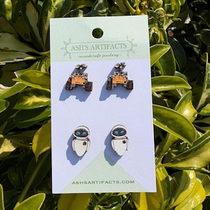 Puede incluir: Un par de pendientes de madera con los personajes Wall-E y Eve de la película de Disney Pixar Wall-E. Los pendientes están en una tarjeta blanca con el texto "ASH'S ARTIFACTS -woodcraft jewelry- ASHSARTIFACTS.COM".
